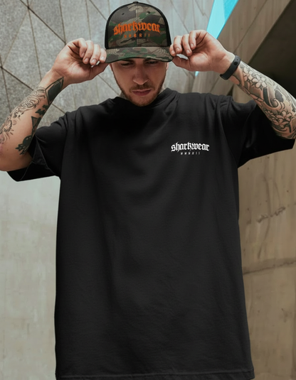 Man wearing a black t-shirt with a logo and a cap with text, standing against a textured wall.